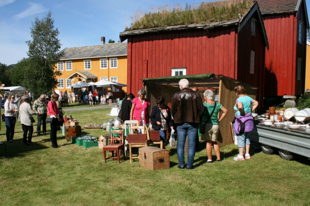 Folkeliv ved sommerarrangement. Mennesker ser på gjenstander foran et rødt stabbur i solskinn.